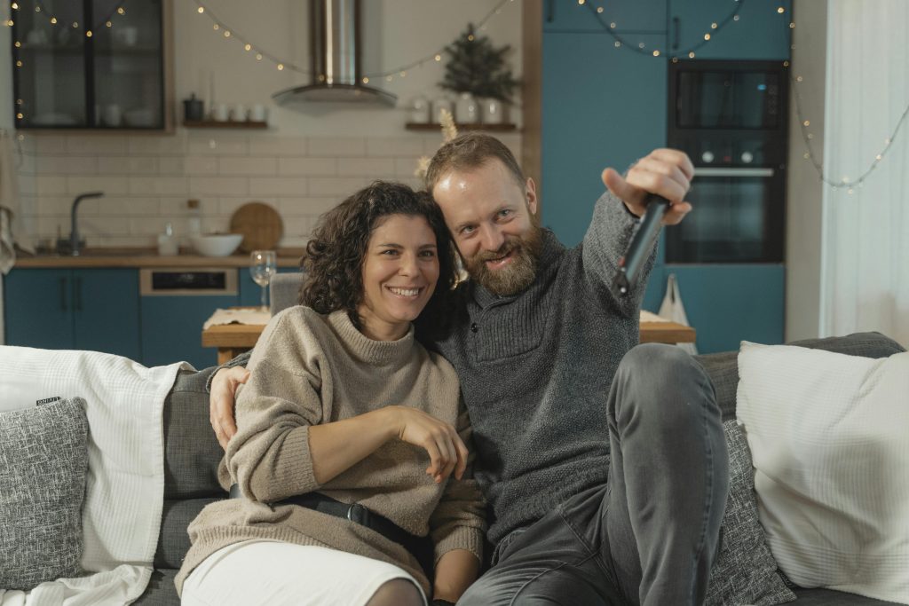 A joyful couple sitting on a couch indoors, watching TV together and smiling.